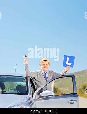 Smiling mature man posing next to his car holding a L sign and car key after having his driver's license Stock Photo