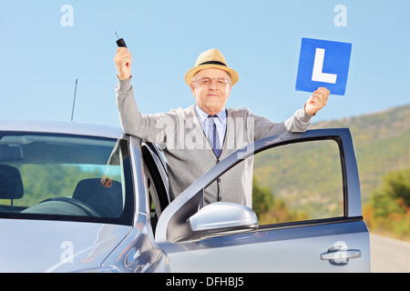 Smiling senior man posing next to his car holding a L sign and car key after having his driver's license Stock Photo