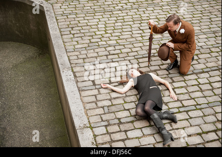 Elevated view of woman lying on cobble stones, with a man in a brown suit kneeling beside her holding an umbrella. Crime scene ? Stock Photo