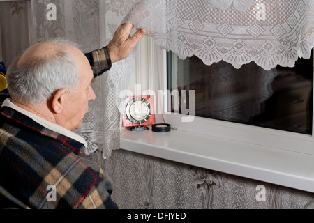 Elderly man peering out through the window into the dark night as he waits for an expected arrival with a clock in front of him Stock Photo