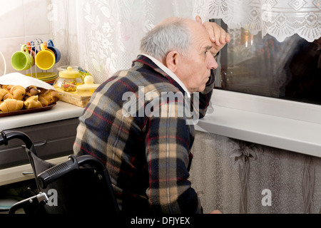 Senior man in a wheelchair peering through a window at night with his hand raised to his forehead Stock Photo