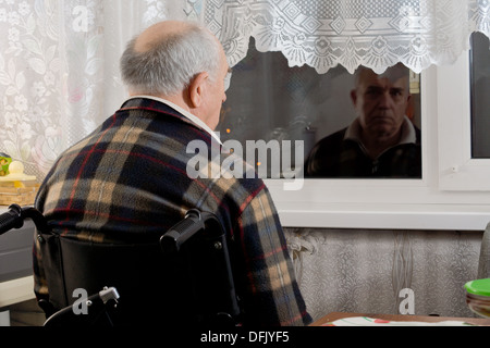 Elderly man in a wheelchair sitting waiting at a window with his back to the camera staring through the glass into the dark night. Stock Photo