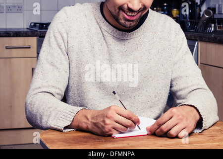 Happy young man sitting at table in kitchen and writing Stock Photo