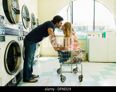 A young man leans over to feel the nose of a beautiful lady who sits on wheeled basket in San Diego coin laundromat.