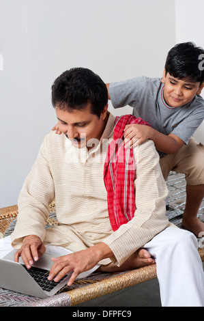 Farmer sitting on a cot and using a laptop with his son behind him Stock Photo