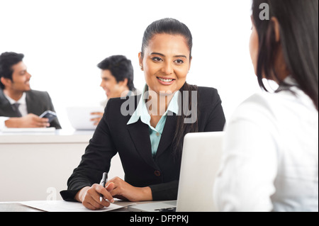 Two businesswomen discussing in an office with their colleagues in the background Stock Photo
