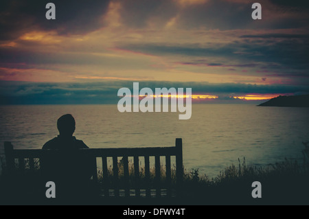 A man sat on a bench looking out at the sunset over the sea Stock Photo