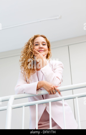 Portrait of confident successful business woman standing in office hallway leaning on railing shot from below Stock Photo
