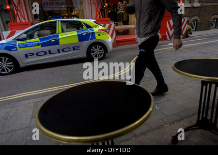 As a Met police car drives past, a young man walks past cafe tables. Stock Photo