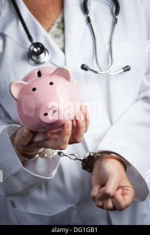 Female Doctor or Nurse In Handcuffs Holding Piggy Bank Wearing Lab Coat and Stethoscope. Stock Photo