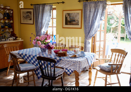 Blue checked cloth on table with stick back chairs in yellow dining room with blue curtains at French windows to the garden Stock Photo