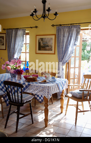 Blue checked cloth on table with stick back chairs in yellow dining room with tiled floor and blue curtains at French windows Stock Photo