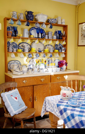 Blue and white china on dresser in yellow dining room with blue cushion on  chair at table with blue checked cloth and floral jug Stock Photo