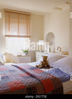 Wooden blind on window in white bedroom with red+blue patchwork quilt and teddy bear on bed with white pillows Stock Photo