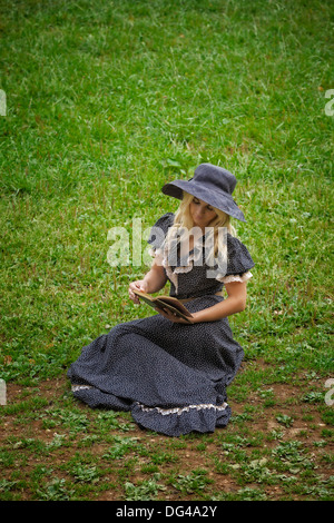 a woman in a romantic dress is sitting on a lawn, reading a book Stock Photo