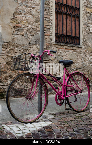 Traditional pink bicycle with a black wire basket tied up to a pole on a cobbled street in Italy. Stock Photo