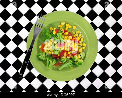 A vegetarian meal of corn, broccoli and red peppers on a green plate with a fork atop a white and black checkerboard tablecloth. Stock Photo
