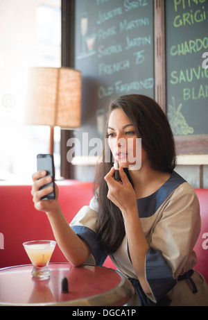 Mid adult women using mobile phone to apply lipstick in restaurant Stock Photo