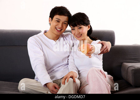 East Asian Couple Embracing, Sitting on the Sofa, Smiling, Looking at the Camera Stock Photo
