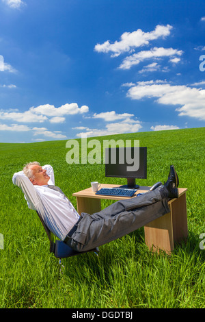 Man or male businessman relaxing feet up at a desk with a computer in a green field drinking tea or coffee Stock Photo
