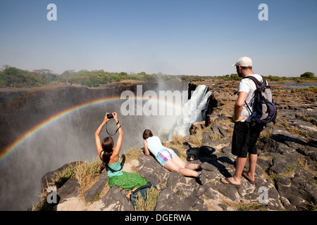 Three tourists looking at the Victoria Falls and rainbow from Livingstone Island, Zambia africa Stock Photo