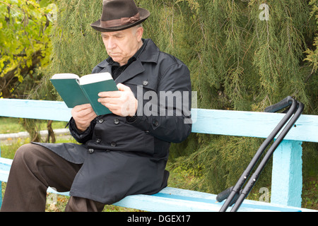 Disabled man on crutches sitting reading in the sun on a rustic wooden bench in the park in his warm overcoat and hat as he enjoys the last sunshine of the autumn. Stock Photo