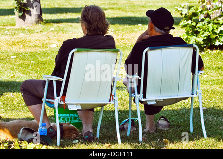 Rear view of couple / man and woman sitting on folding chairs in public park - France. Stock Photo