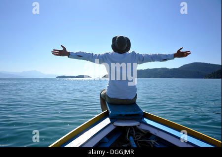 Young man sitting on the bow of a fishing boat with his arms wide open, Bay of Paraty or Parati, State of Rio de Janeiro, Brazil Stock Photo