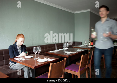 Young woman using cell phone in restaurant, waiter walking past Stock Photo