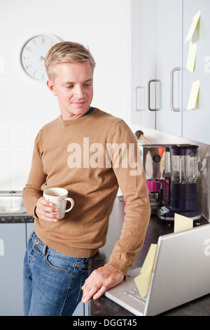 Young man in kitchen with coffee cup and laptop Stock Photo