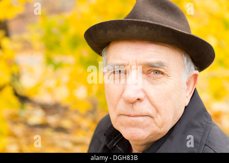 Close up portrait of an elderly man looking at the camera with a serious expression wearing a stylish hat while enjoying time outdoors in a colourful autumn garden. Stock Photo