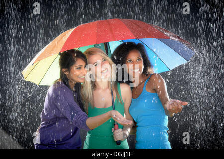 Three smiling young women of Cuban, Indian and German descent, standing together under a colourful rainbow umbrella enjoying the Stock Photo