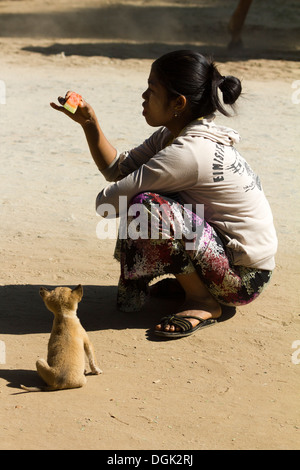 A woman eating watermelon in the middle of the road whilst a puppy watches at Mingun in Myanmar. Stock Photo