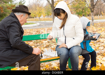 Attractive woman sitting on a park bench in the cold autumn weather playing chess with an elderly man while her young son amuses himself on a tablet computer behind her. Stock Photo