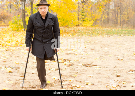 Lonely one-legged senior Caucasian man walking with crutches in an Autumn day in the park, with withered foliage in the background. Stock Photo