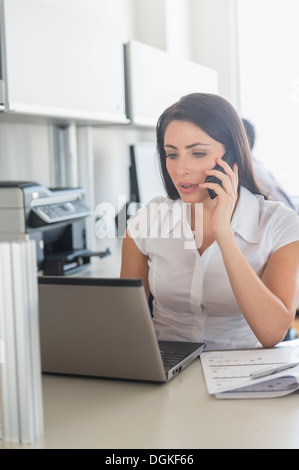 Woman working on laptop and talking on cell phone in office, man in background Stock Photo