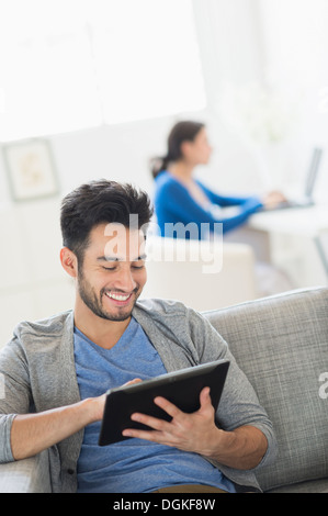 Man sitting on sofa using digital tablet at home, woman in background Stock Photo