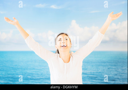 Portrait of woman with arms raised in front of backdrop with sea and sky Stock Photo