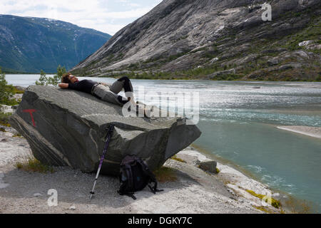 Woman wearing hiking clothing lying on a prominent boulder at the glacial lake of Nigardsbrevatnet, Jostedal Stock Photo