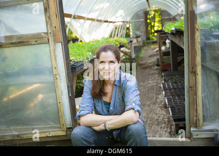 On the farm. A woman sitting resting at the door of a glasshouse with benches full of young seedling plants. Stock Photo