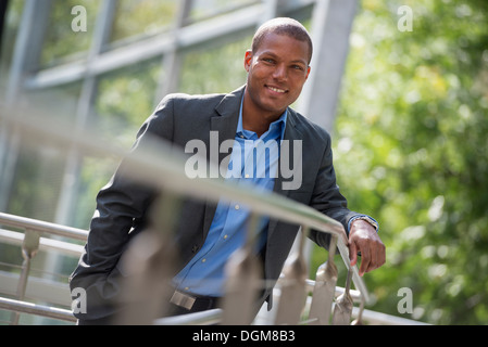 A young man in a jacket and open collared blue shirt, leaning on a railing in a city park in summer. Looking at the camera. Stock Photo