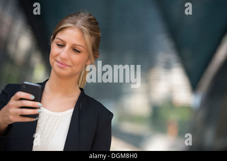 A young blonde businesswoman on a New York city street. Wearing a black jacket. Using a smart phone.