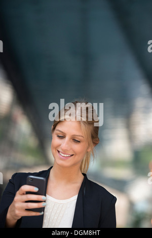 A young blonde businesswoman on a New York city street. Wearing a black jacket. Using a smart phone.