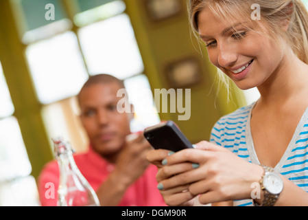 A couple in a city coffee shop. A woman sitting down checking a smart phone. A man in the background.