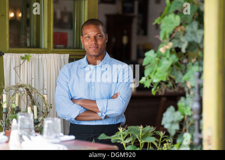 A man standing by the open window of a cafe or bistro, looking out with arms folded. Stock Photo
