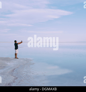 A man standing at edge of the flooded Bonneville Salt Flats at dusk, taking a photograph with a tablet device, near Wendover. Stock Photo