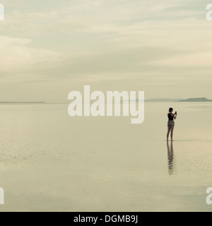 A woman standing on the flooded Bonneville Salt Flats, taking a photograph with a smart phone. Stock Photo