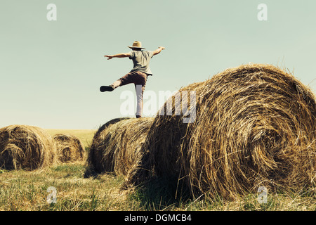 Back view of a man balancing on one leg on top of a hay bale.