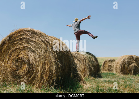 Back view of a man balancing on one leg on top of a hay bale.