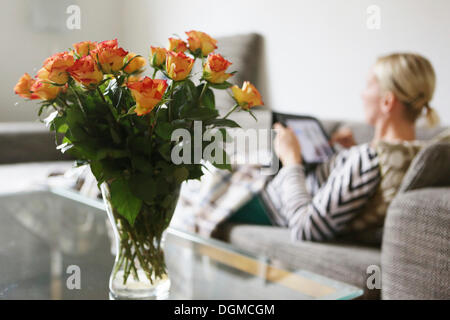 Bouquet of roses in a living room, a young woman lying on the sofa with a tablet computer, at back Stock Photo